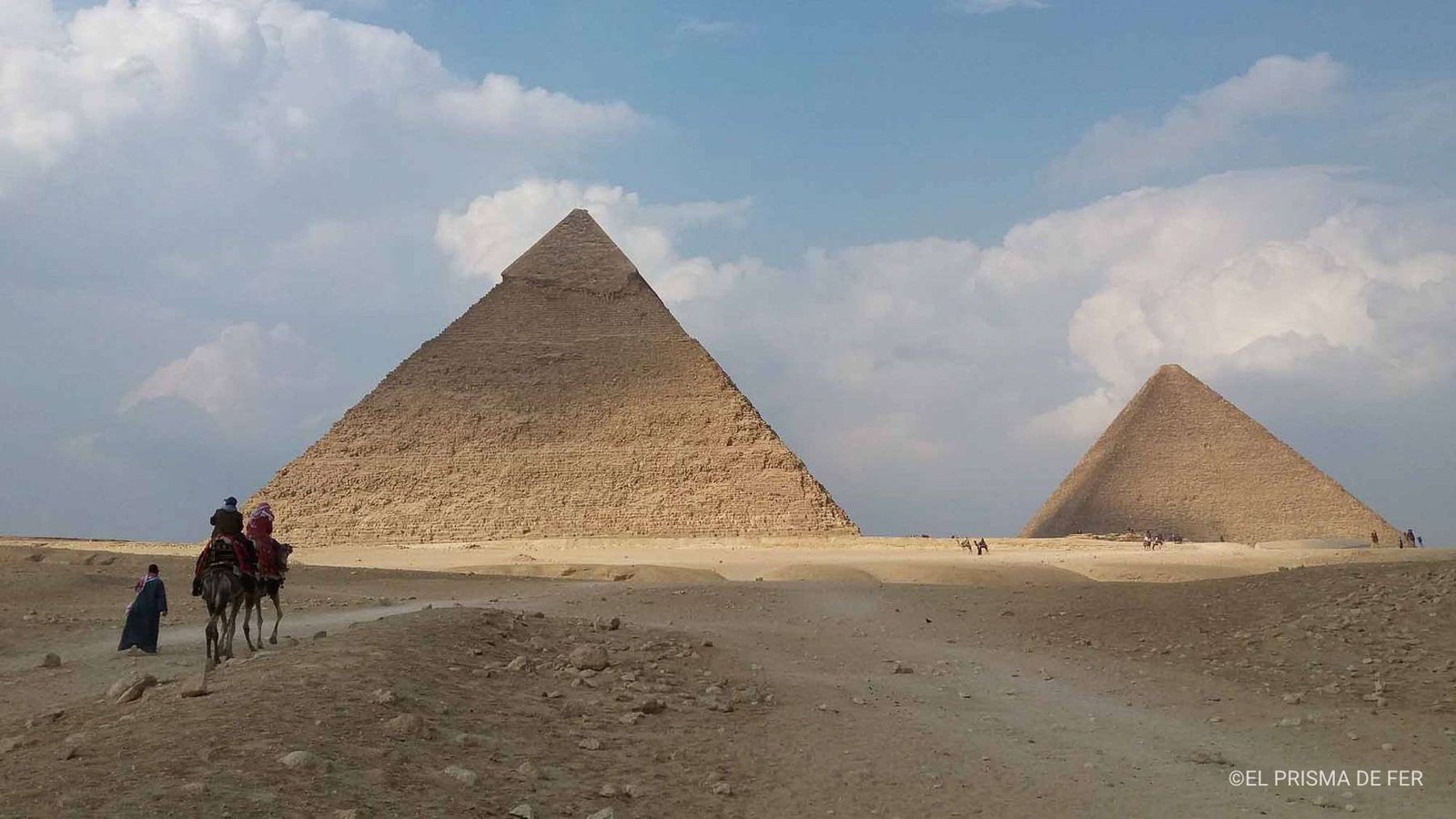 Vista icónica das Pirâmides de Gizé sob um céu azul brilhante, com viajantes a montar camelos pelo deserto