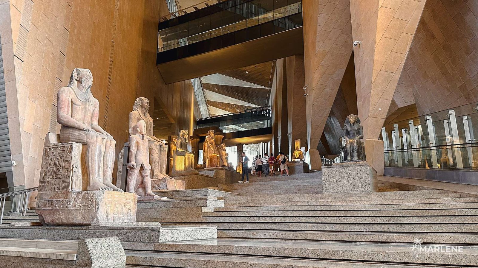 Visitantes a percorrer a escadaria principal do Grande Museu Egípcio, observando as grandiosas esculturas expostas sob a luz do átrio.