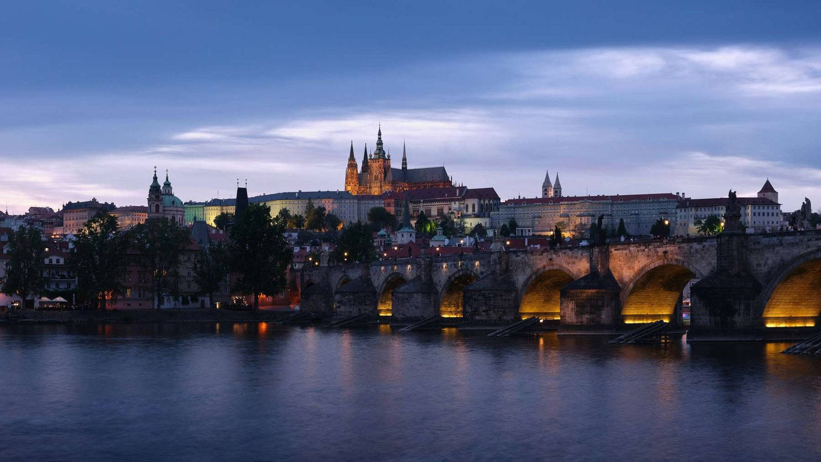 A Ponte Carlos e o Castelo de Praga refletidos no rio Moldava ao entardecer.